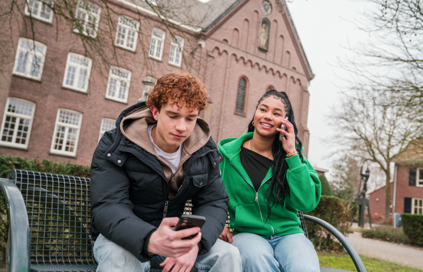 jongen en meisje zitten op bankje, meisje telefoneert en jongen kijkt op zijn smartphone