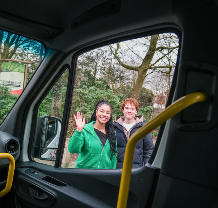 Twee personen zwaaien door raam, foto is vanuit bus gemaakt.